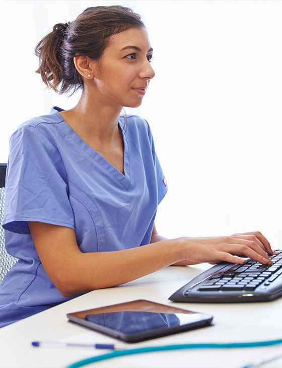 young female nurse in blue scrubs using a computer keyboard with tablet and pen on desk focused on digital healthcare solutions